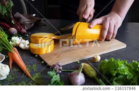 Close up of young woman hands cutting zucchini on wooden cutting board at home. Young woman preparing healthy food. 116157846