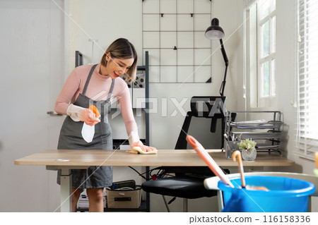 Young Woman Cleaning Desk in Modern Home Office with Cleaning Supplies and Natural Light 116158336