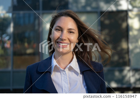 Portrait of an attractive young adult woman in a blue suit standing against the background of an office building, smiling and looking at the camera close-up 116158947