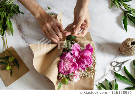 A female florist makes a bouquet of peony flowers wrapped in kraft paper on her desktop. View from above. High quality photos 116158985