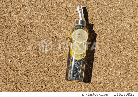 Transparent glass bottle of refreshing water with lemon and blueberries in sunny day on outdoor beige sports ground. Concept water balance for heat season for workout. View from above. Copy space. Transparent glass bottle of refreshing water with lemon and blueberries in sunny day on outdoor beige sports ground. Concept water balance for heat season for workout. View from above. Copy space. 116159822