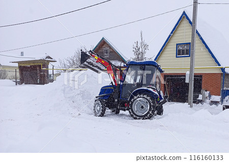 Mini tractor with a raised bucket spreads snow while clearing snow on a winter day. City road services for cleaning and landscaping. Mechanization of labor as an increase in production efficiency. 116160133