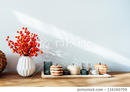 Cozy autumn home interior - various decorative wicker pumpkins, candles, seasonal flowers in vase on the wooden console with white wall background. Scandinavian minimalist hygge home fall decor. 116160709