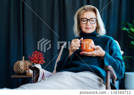 Middle aged woman relaxing with pumpkin shaped cup of hot drink in scandy style hygge interior home with fall mood decor. Lady dreaming, enjoy calm mood without stress, well being alone. Cozy autumn. Middle aged woman relaxing with pumpkin shaped cup of hot drink in scandy style hygge interior home with fall mood decor. Lady dreaming, enjoy calm mood without stress, well being alone. Cozy autumn. 116160712