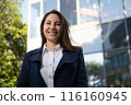 Portrait of a happy caucasian brunette woman in a blue suit stands against the background of a business building in the park and smiles 116160945