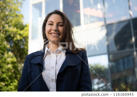 Portrait of a happy caucasian brunette woman in a blue suit stands against the background of a business building in the park and smiles 116160945