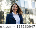 Portrait of a happy caucasian brunette woman in a blue suit stands against the background of a business building and smiles 116160947