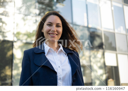 Portrait of a happy caucasian brunette woman in a blue suit stands against the background of a business building and smiles Portrait of a happy caucasian brunette woman in a blue suit stands against the background of a business building and smiles 116160947