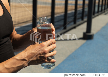 Athletes man holding glass bottle of water on outdoor sports ground. Concept water balance for hot season for active workout. Close up. Copy space. Athletes man holding glass bottle of water on outdoor sports ground. Concept water balance for hot season for active workout. Close up. Copy space. 116163195