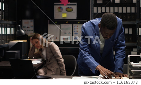 African american male detective diligently analyzes evidence and classified documents to solve a case. Frustrated private investigator stands and reviews confidential clues and witness statements. 116165378