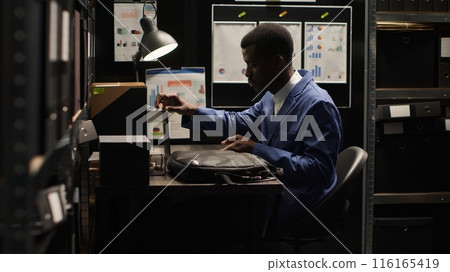 African American investigator arrives office in adorned with crime scene photos and marked-up maps, carrying laptop bag. Placing laptop on table, detective meticulously reviews evidence files. 116165419
