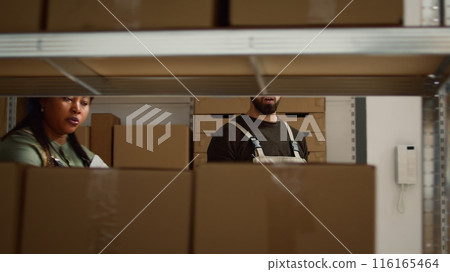 Team leader and assistant walking through shelves preparing warehouse orders for delivery, using devices to verify shipping informations, crosschecking online data and scanning wares labels Team leader and assistant walking through shelves preparing warehouse orders for delivery, using devices to verify shipping informations, crosschecking online data and scanning wares labels 116165464