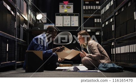 In office space, male private detective examines case files and reads witness statements from folder. Sitting on floor, two investigators analyze crucial evidence for criminal investigation. Zoom-out. In office space, male private detective examines case files and reads witness statements from folder. Sitting on floor, two investigators analyze crucial evidence for criminal investigation. Zoom-out. 116165470
