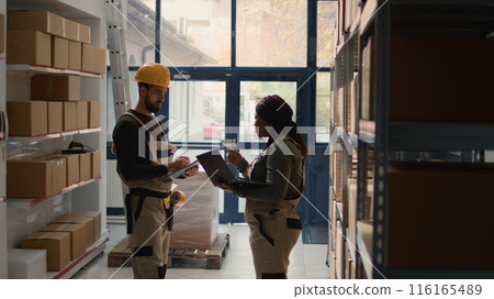 Warehouse chief supply chain officers inputing updated products info on online shopping website, walking through storeroom aisles inspecting labels on wares, crosschecking with inventory list 116165489