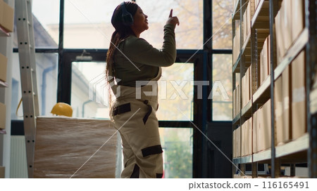 Happy warehouse manager listening music enjoying herself while examining merchandise. Cheerful supervisor wearing heaphones and dancing around while walking through storage facility rows Happy warehouse manager listening music enjoying herself while examining merchandise. Cheerful supervisor wearing heaphones and dancing around while walking through storage facility rows 116165491