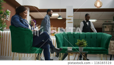 Hotel guest verifying webpage at hotel, waiting for accommodation at holiday resort. African american woman using laptop to review files and check social media before seeing room. Hotel guest verifying webpage at hotel, waiting for accommodation at holiday resort. African american woman using laptop to review files and check social media before seeing room. 116165519