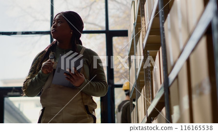 BIPOC warehouse stocker performing inventory survey, keeping quality standards high. Productive expert in distribution center preparing to ship parcels, crosschecking shippment info 116165538