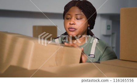 African american employee in warehouse handling cardboard boxes full of products ready to be shipped to customers. Worker in storage room placing parcels on cabinet shelves 116165556