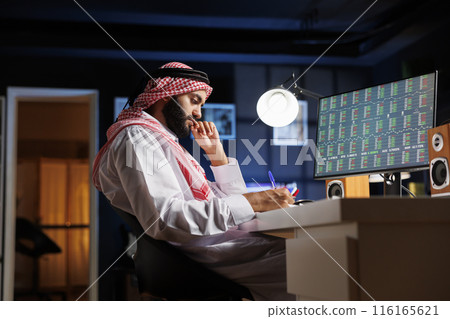 Middle Eastern guy sat at his workstation, intently researching and taking notes from his desktop computer. This image displays an Arab man with a notepad who is brainstorming and using a computer. 116165621