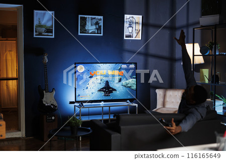 African american man celebrates tournament success, playing online video gaming competition and winning. Young player having fun with virtual long range weapons, winning space shooter contest. African american man celebrates tournament success, playing online video gaming competition and winning. Young player having fun with virtual long range weapons, winning space shooter contest. 116165649