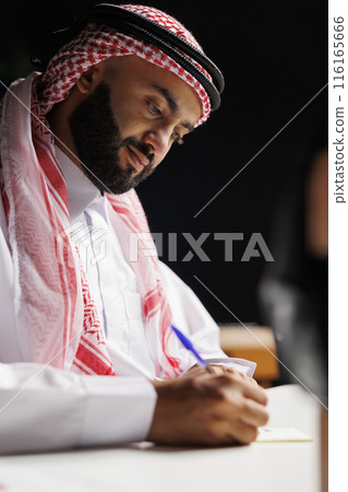 The image portrays an attentive Middle Eastern guy in traditional clothing working at a table with a notepad. An Islamic man is shown writing research at his desk at home in a close-up shot. 116165666