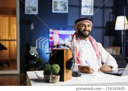Portrait shot of Muslim man freelancer using laptop to browse the internet while carrying a pen. Side-view of Arab guy looking at camera, displaying his professional profile in contemporary workplace. 116165685
