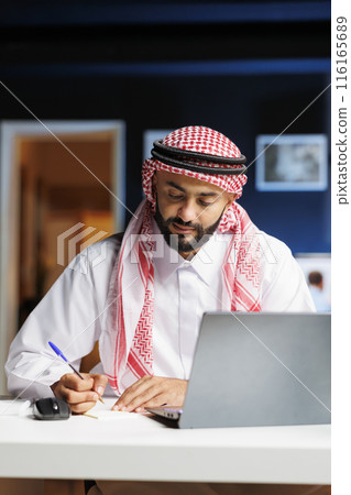 An Arab businessman, absorbed in work, diligently browses digital information on office desk laptop. He takes notes while conducting internet research, demonstrating his professionalism and attention. 116165689