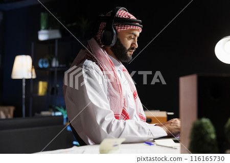 Muslim freelancer works diligently and uses modern technology from desk. Engaged in online research and communication, he wears traditional attire and listens attentively through wireless headphones. Muslim freelancer works diligently and uses modern technology from desk. Engaged in online research and communication, he wears traditional attire and listens attentively through wireless headphones. 116165730