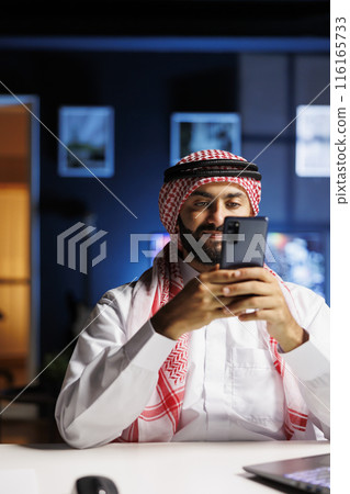 Holding a digital mobile device is a young man in Arabian clothing sitting at a desk. Muslim guy Holding a digital mobile device is a young man in Arabian clothing sitting at a desk. Muslim guy 116165733