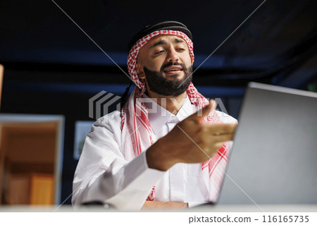 Young businessman representing success in the digital era, wearing Arabian apparel, using a digital laptop. Muslim guy conversing confidently on his personal computer at his home office desk. Young businessman representing success in the digital era, wearing Arabian apparel, using a digital laptop. Muslim guy conversing confidently on his personal computer at his home office desk. 116165735