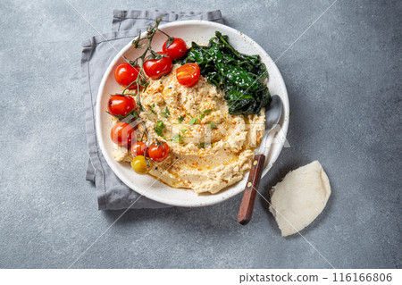 A delicious plate of creamy hummus garnished with fresh tomatoes, leafy greens, and served with pita bread 116166806