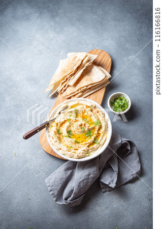 Bowl of creamy hummus garnished with olive oil and herbs is served with pita bread on a wooden board. A small bowl of garnish is placed beside the bread on a slate surface 116166816