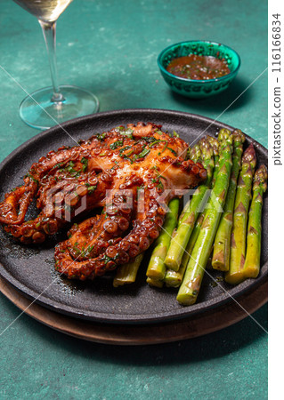 Grilled octopus with asparagus served on a black plate, highlighting vibrant colors and textures against a turquoise background. Grilled octopus with asparagus served on a black plate, highlighting vibrant colors and textures against a turquoise background. 116166834