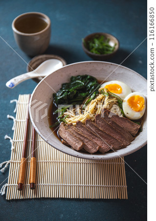 Bowl of ramen with slices of meat, soft-boiled eggs, spinach, and noodles in rich broth, served with chopsticks on a bamboo mat 116166850