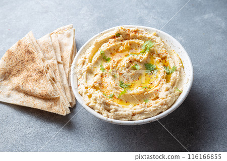 Bowl of creamy hummus garnished with olive oil and herbs is served with pita bread on a wooden board. A small bowl of garnish is placed beside the bread on a slate surface Bowl of creamy hummus garnished with olive oil and herbs is served with pita bread on a wooden board. A small bowl of garnish is placed beside the bread on a slate surface 116166855