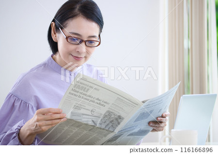 A middle-aged woman wearing reading glasses and reading an English newspaper 116166896