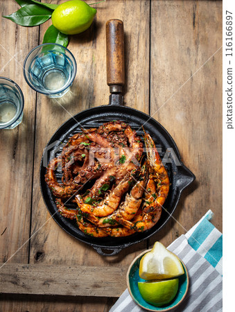 Grilled shrimps and octopus on a black grill pan on a rustic wooden table, with limes 116166897
