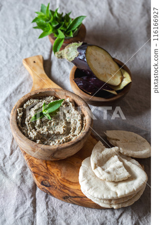 Wooden bowl of Jewish baba ganoush garnished with eggplant and mint leaves sits on a wooden board. Surrounding the bowl are soft pita bread slices and a bowl of fresh eggplant slices. Wooden bowl of Jewish baba ganoush garnished with eggplant and mint leaves sits on a wooden board. Surrounding the bowl are soft pita bread slices and a bowl of fresh eggplant slices. 116166927