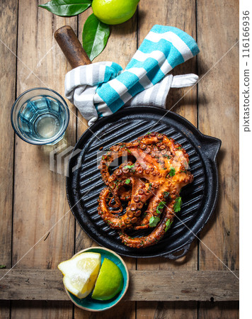 Grilled octopus on a black grill pan on a rustic wooden table, with limes, a blue-striped cloth 116166936