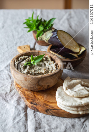Wooden bowl of Jewish baba ganoush garnished with eggplant and mint leaves sits on a wooden board. Surrounding the bowl are soft pita bread slices and a bowl of fresh eggplant slices. Wooden bowl of Jewish baba ganoush garnished with eggplant and mint leaves sits on a wooden board. Surrounding the bowl are soft pita bread slices and a bowl of fresh eggplant slices. 116167011