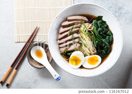 A bowl of ramen with slices of beef, soft-boiled egg halves, and green vegetables. The meal is garnished with green onions 116167012