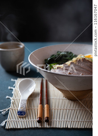 Bowl of ramen with slices of meat, soft-boiled eggs, spinach, and noodles in rich broth, served with chopsticks on a bamboo mat 116167047