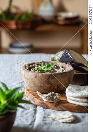 Wooden bowl of Jewish baba ganoush garnished with eggplant and mint leaves sits on a wooden board. Surrounding the bowl are soft pita bread slices and a bowl of fresh eggplant slices. Wooden bowl of Jewish baba ganoush garnished with eggplant and mint leaves sits on a wooden board. Surrounding the bowl are soft pita bread slices and a bowl of fresh eggplant slices. 116167054