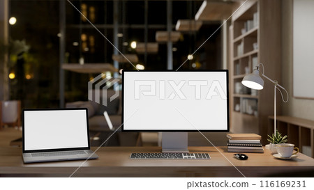 A modern office workspace at night features a laptop and computer mockup on a wooden table. 116169231