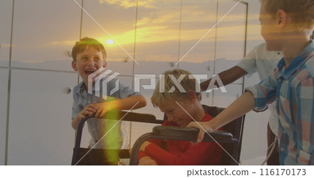 Image of sunset landscape over happy diverse schoolboys pushing friend in wheelchair 116170173