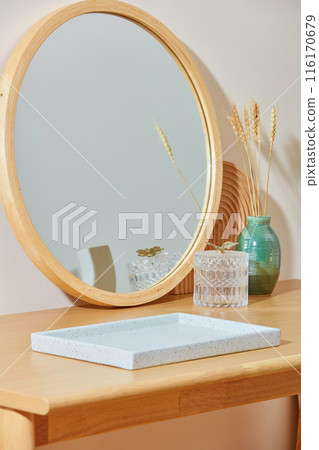 A large empty tray in white color displayed on a wooden table, behind it is decorated with a big mirror, a glass pot and a flower vase. Photo has diagonal angle, copy space for text 116170679