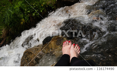 Feet with red toenails near mountain stream with lush greenery. 116174490