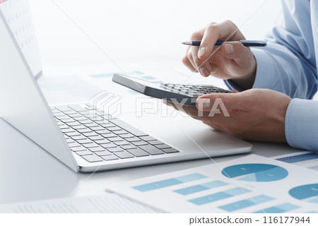 A man calculating taxes using a calculator and a laptop. Tax returns, year-end adjustments, and other tax returns. 116177944