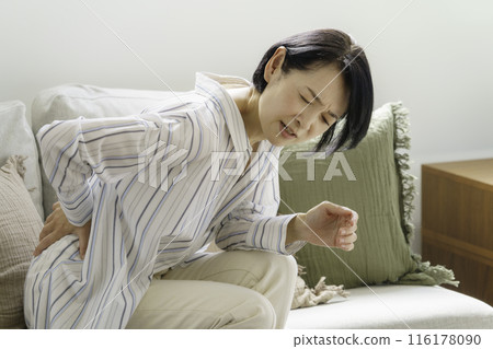 Middle-aged woman sitting on sofa holding her back, back pain Middle-aged woman sitting on sofa holding her back, back pain 116178090