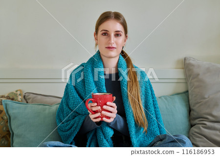 Young woman warming under blanket with mug of hot drink sitting on couch at home Young woman warming under blanket with mug of hot drink sitting on couch at home 116186953
